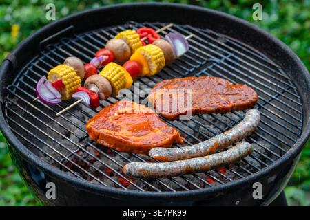 Steaks, saucisses et brochettes de légumes colorées grillant sur un barbecue au charbon de bois. Scène de cuisine extérieure avec maïs, champignons, oignon et poivrons. Banque D'Images