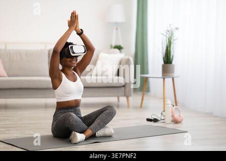 Femme noire avec des vêtements de sport bouclés et casque VR méditant sur tapis dans le salon à la maison Banque D'Images