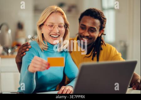 Souriant homme de race mixte avec serrures et petite amie caucasienne tenant la carte de crédit assis à table avec ordinateur portable à la maison Banque D'Images