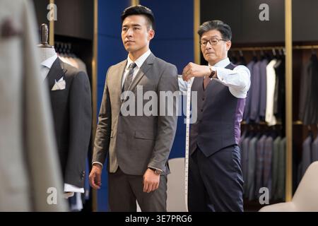 Concepteurs de mode masculins chinois seniors prenant la mesure du client masculin chinois tout en se tenant dans la boutique de mode Banque D'Images