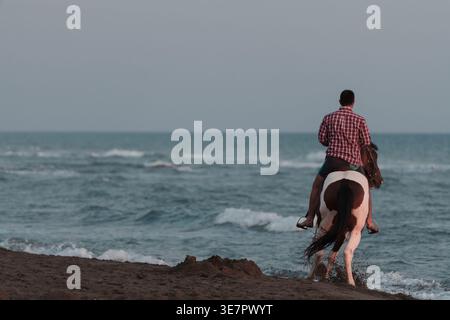 Homme en chemise rouge à carreaux à manches longues chevauchant un cheval sur la plage au coucher du soleil Banque D'Images