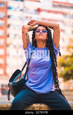 Femme hispanique avec de longues grandes torsions portant chemise violette et lunettes assis sur une clôture en béton avec les bras sur le dessus de la tête dans la rue de la ville Banque D'Images