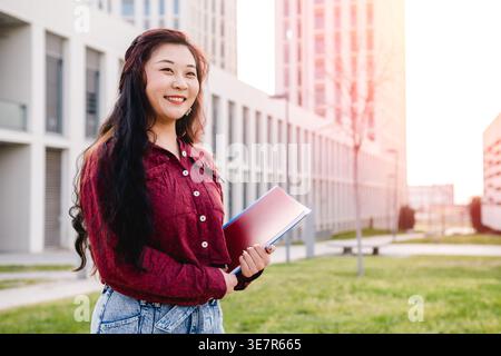 Souriante étudiante asiatique tenant des cahiers et regardant loin tout en se tenant près du bâtiment de l'école sur le campus de l'université Banque D'Images