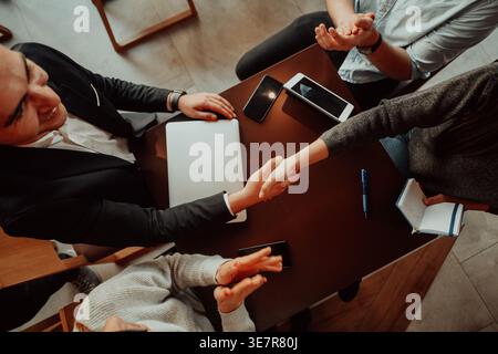 Groupe diversifié de gens d'affaires serrant la main tout en étant assis à table avec ordinateur portable et smLifestylephone dans le café Banque D'Images