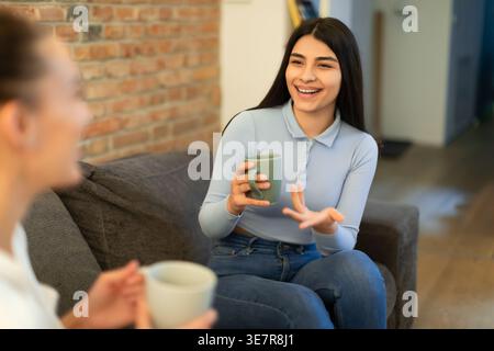 Femme du moyen-Orient tenant une tasse de café parlant à une amie féminine tout en étant assise sur le canapé dans le salon à la maison Banque D'Images