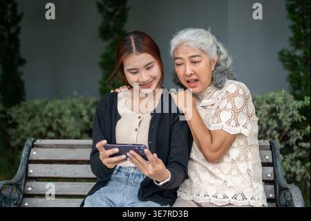 Femme thaïlandaise ayant du plaisir à partager smartphone avec sa grand-mère tout en étant assis sur un banc en bois dans le parc pendant la journée Banque D'Images