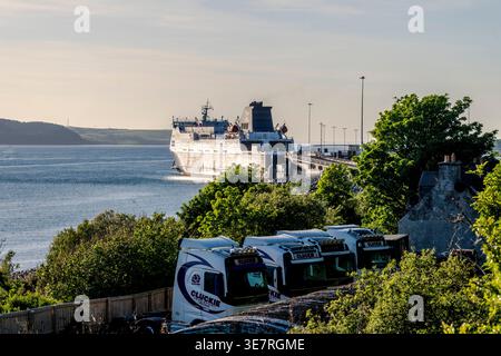 Un grand ferry accoste à Cairnryan, avec des camions garés à côté d'une végétation luxuriante et d'une eau calme. Banque D'Images