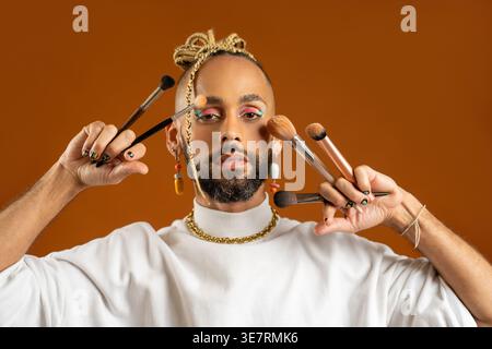 Homme gay brésilien sérieux avec des cheveux tressés portant un pull blanc à col roulé avec maquillage coloré des yeux tenant des pinceaux de maquillage debout contre Ora... Banque D'Images