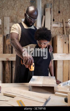 Père noir dans le tablier portant des lunettes debout derrière le fils l'aidant à percer le bois dans l'atelier Banque D'Images