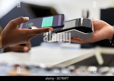 Mains d'une femme noire payant à l'aide d'un smartphone à un caissier masculin caucasien tenant un terminal POS tout en se tenant debout au-dessus du comptoir dans un magasin de vêtements Banque D'Images