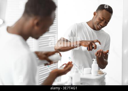 Homme noir en chemise blanche mettant de la mousse à raser sur sa main tout en se tenant devant le miroir dans la salle de bain à la maison Banque D'Images