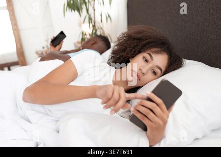 Femme noire avec les cheveux bouclés à l'aide d'un smartphone tout en étant couché sur le lit contre son petit ami dans la chambre à coucher à la maison Banque D'Images
