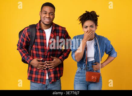 Intoxication alimentaire en voyage de vacances. Couple de touristes noirs ayant des nausées et des maux d'estomac souffrant de douleurs abdominales debout sur jaune Studio dos... Banque D'Images