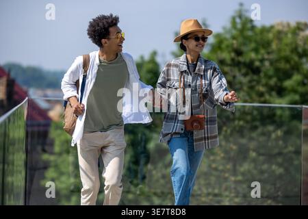 Couple de touristes noirs portant chapeau et lunettes de soleil tenant la main tout en courant près de la clôture de verre dans le parc le jour ensoleillé Banque D'Images