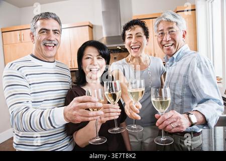 Groupe diversifié d'amis seniors grillant des verres à vin tout en se tenant debout dans la cuisine à la maison Banque D'Images