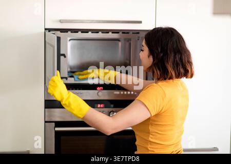 Adolescente hispanique portant des gants en caoutchouc en utilisant un chiffon dans le micro-ondes de nettoyage tout en se tenant debout dans la cuisine à la maison Banque D'Images