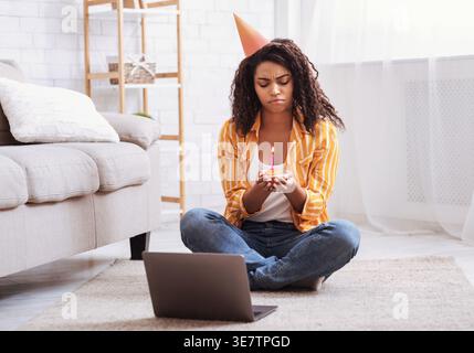 Triste femme noire portant chapeau de fête tenant cupcake avec bougie tout en étant assis sur le tapis de sol avec ordinateur portable dans le salon à la maison Banque D'Images