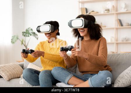 Amies noires portant VR expérimentant la réalité virtuelle en utilisant le contrôleur de jeu s'amusant tout en étant assis sur le canapé dans le salon à la maison Banque D'Images
