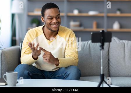 Homme noir souriant faisant du streaming en direct à l'aide d'un smartphone sur trépied donnant une leçon en ligne assis sur un canapé dans le salon à la maison Banque D'Images