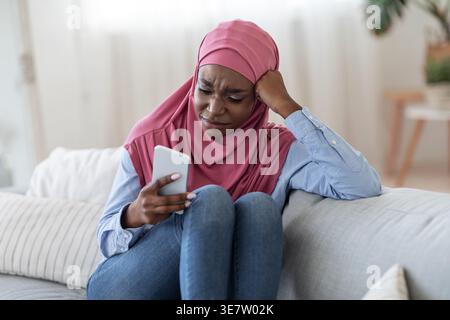 Femme noire portant le hijab à l'aide d'un smartphone tout en étant assis sur un canapé dans le salon à la maison Banque D'Images