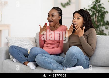 Amies noires effrayées regardant un film ensemble tout en étant assis sur un canapé dans le salon à la maison Banque D'Images