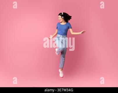 Femme asiatique portant un casque sans fil écoutant de la musique tout en sautant et dansant avec les yeux fermés sur fond rose en studio Banque D'Images