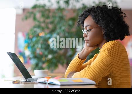 Femme noire avec des cheveux bouclés portant des lunettes penchant le visage sur la main regardant l'ordinateur portable assis à table avec ordinateur portable et tasse à café dans le café Banque D'Images