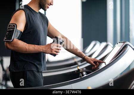 Image recadrée d'un homme noir avec une barbe portant un brassard tenant une bouteille shaker et sélectionnant le mode sur le tapis roulant avant de faire du jogging dans la salle de gym Banque D'Images