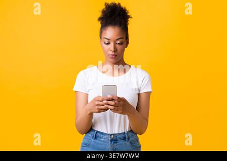Femme noire utilisant un smartphone debout sur fond jaune dans le studio Banque D'Images
