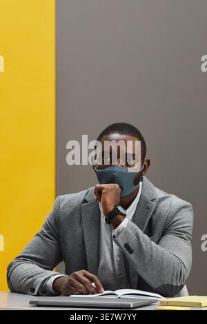 Homme d'affaires afro-américain en costume gris portant un masque facial assis devant un fond gris et jaune dans le studio Banque D'Images