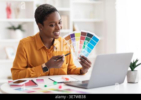 Designer féminin noir avec les cheveux courts ayant la vidéo conférence sur ordinateur portable et tenant des palettes de couleurs tout en étant assis à table avec smartphone et noteb... Banque D'Images