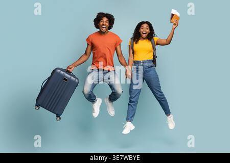 Joyeux homme noir avec la bouche ouverte portant valise tenant la main avec la petite amie tenant les passeports et les billets tout en sautant contre le backgro bleu... Banque D'Images