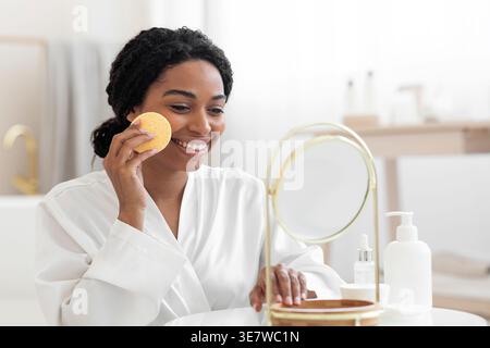 Femme noire portant peignoir nettoyant le visage à l'aide d'une éponge regardant le miroir tout en étant assis à table dans la salle de bain à la maison Banque D'Images