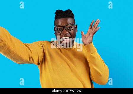 Joyeux homme noir avec la bouche ouverte portant des lunettes gestuelles main prenant selfie tout en se tenant debout sur fond bleu en studio Banque D'Images