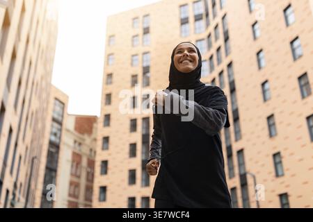 Femme musulmane du moyen-Orient souriante avec tracker de fitness portant hijab et vêtements de sport courant dans la rue pendant la journée Banque D'Images