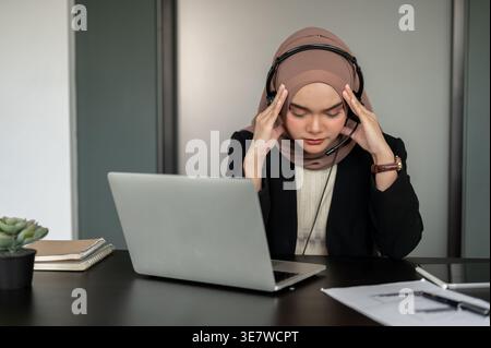 Service client asiatique stressé portant un hijab et un casque touchant les tempes tout en étant assis à table avec un ordinateur portable au bureau Banque D'Images