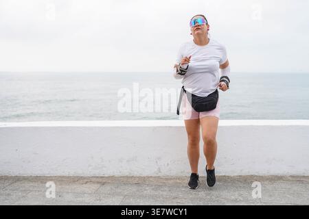 Femme hispanique portant des poids au poignet et des lunettes de soleil jogging par la mer en ville Banque D'Images