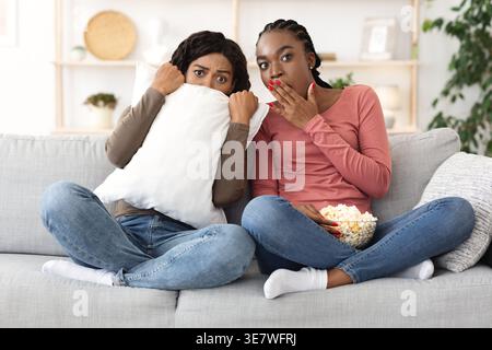 Amies noires effrayées regardant un film ensemble tout en étant assis sur un canapé dans le salon à la maison Banque D'Images