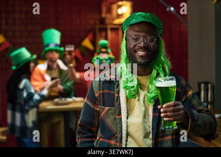 Homme noir en casquette verte et perruque tenant un verre de boisson verte tout en célébrant la Saint Patrick avec des amis au bar Banque D'Images