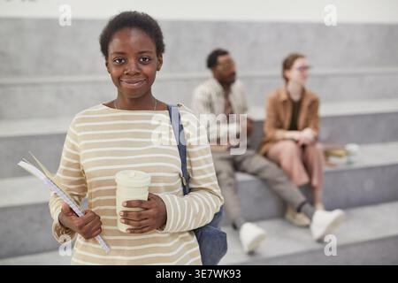 Étudiante universitaire noire avec sac à dos tenant une tasse à café jetable et un cahier debout devant les étudiants dans le hall du collège Banque D'Images