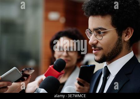 Homme d'affaires de race mixte en costume et lunettes parlant aux journalistes donnant une interview à la conférence d'affaires tout en se tenant debout à l'extérieur du bureau bui... Banque D'Images