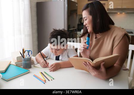 Mère asiatique souriante avec fils de race mixte faisant des devoirs à la maison assis sur le devant de la table à la maison pendant la journée Banque D'Images