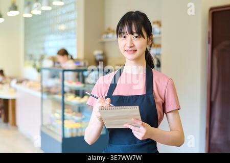 Serveuse asiatique dans un tablier noir debout avec bloc-notes prenant les commandes des clients à l'intérieur du restaurant Banque D'Images