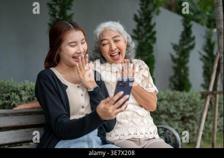 Femme thaïlandaise ayant du plaisir à partager smartphone avec sa grand-mère tout en étant assise sur un banc en bois dans le jardin pendant la journée Banque D'Images