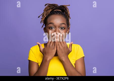Femme noire avec des serrures portant une chemise jaune couvrant sa bouche avec les mains debout sur fond violet dans le studio Banque D'Images