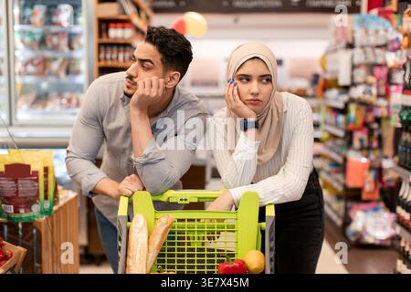 Couple du moyen-Orient penchant le menton sur leurs mains poussant le chariot tout en marchant dans le supermarché Banque D'Images