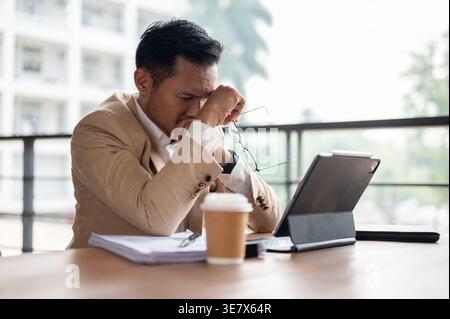 Homme d'affaires asiatique frustré penché le visage sur les deux mains assis à la table en bois avec des documents, tablette et café sur le balcon de son appartement Banque D'Images