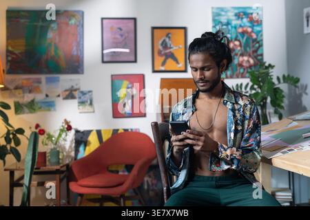 Artiste masculin indien sérieux à l'aide d'un smartphone assis sur une chaise contre ses diverses peintures en arrière-plan dans le home studio pendant la journée Banque D'Images