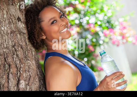 Joyeuse femme noire avec afro bouclé portant débardeur bleu appuyé contre l'arbre et tenant une bouteille d'eau tout en se tenant dans le parc pendant la journée Banque D'Images