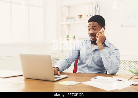 Homme d'affaires noir surpris ayant un appel téléphonique et travaillant sur ordinateur portable assis au bureau en bois dans le bureau Banque D'Images